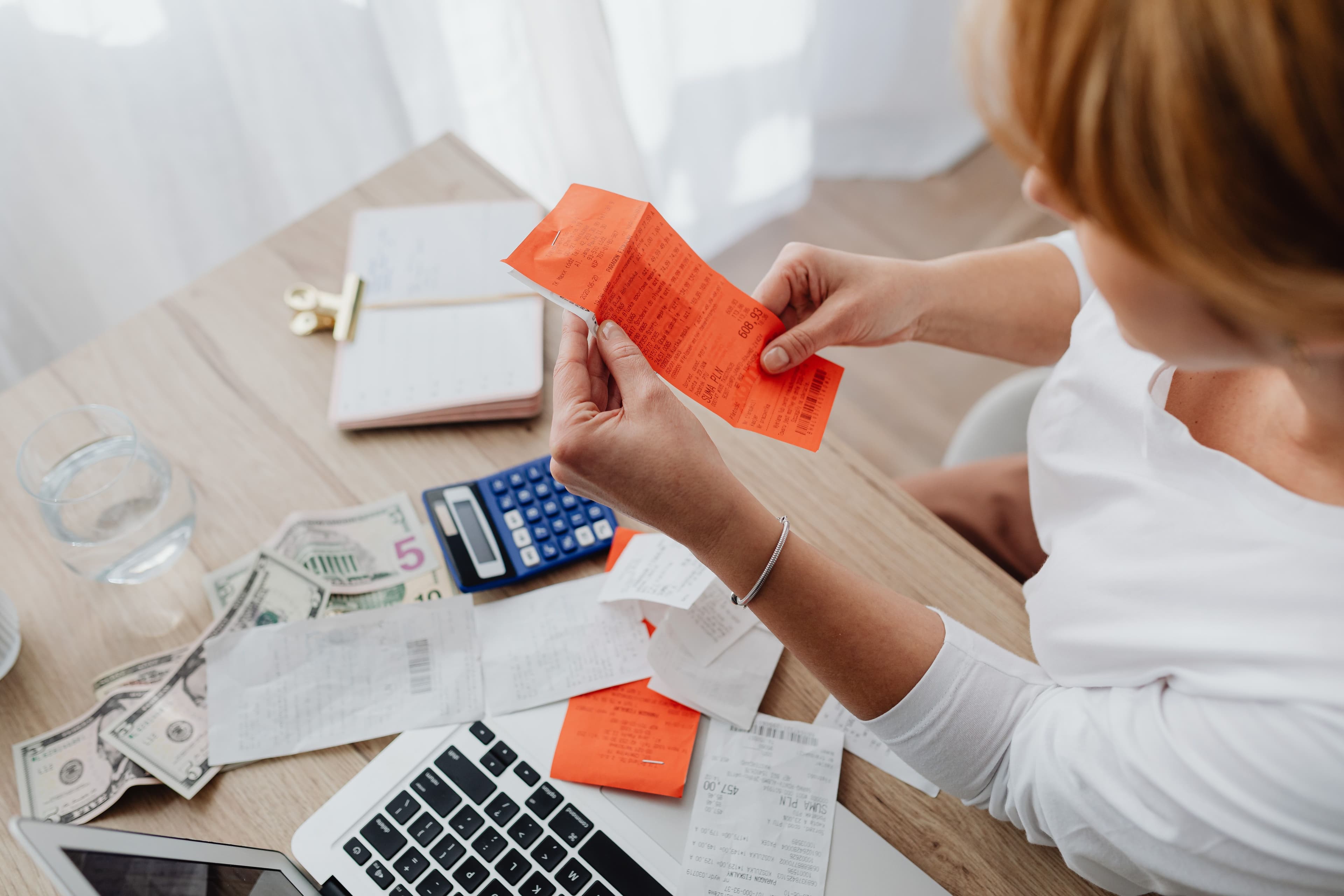 A woman sits at a wooden desk sorting through a stack of paper receipts, with a calculator, cash and a laptop spread out in front of her.